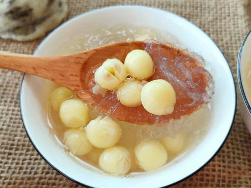 Chè hạt sen với nước trong, hạt sen mềm mịn, được múc trong thìa gỗ, đặt trong bát trắng trên nền vải.
