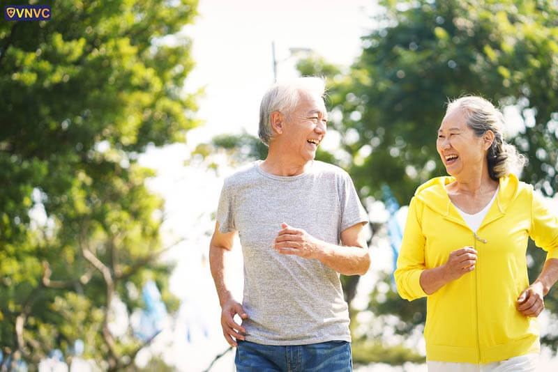 Hai người cao tuổi đang đi bộ và cười vui vẻ trong công viên, thể hiện lối sống lành mạnh và hạnh phúc.