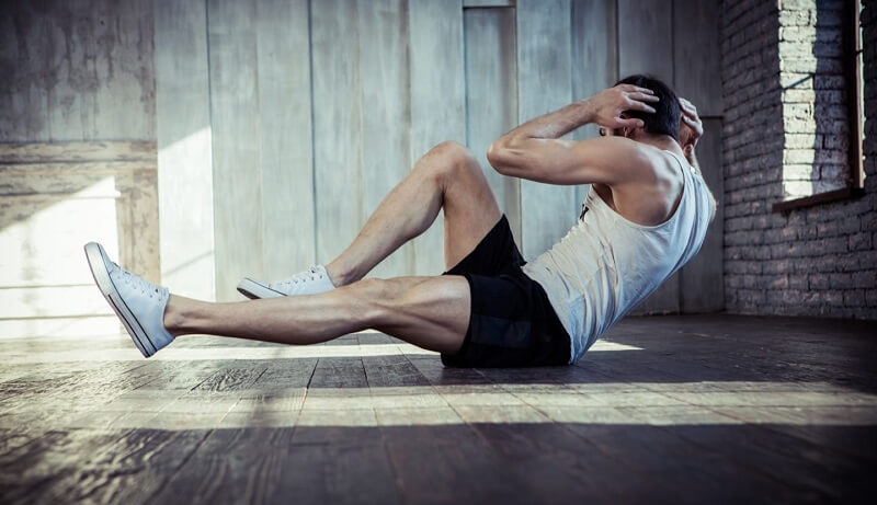Người đàn ông tập thể dục trong phòng gym, mặc áo ba lỗ trắng và quần đen, thực hiện động tác gập bụng trên sàn gỗ.